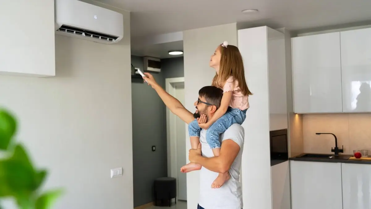 Père et fille ajustant un climatiseur avec une télécommande dans une maison, utilisant une pompe à chaleur air-air pour chauffer ou refroidir l'air intérieur.